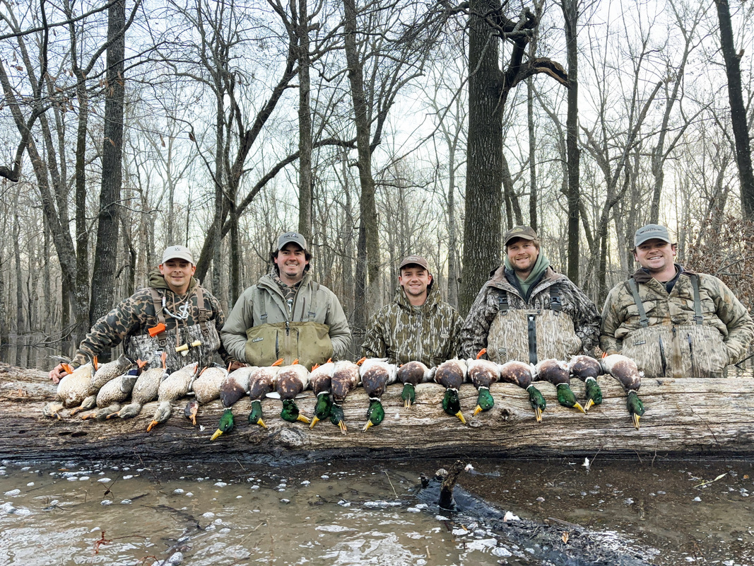 A group of smiling young men in hunting garb with a collection of hunted ducks lined up along a felled tree. 